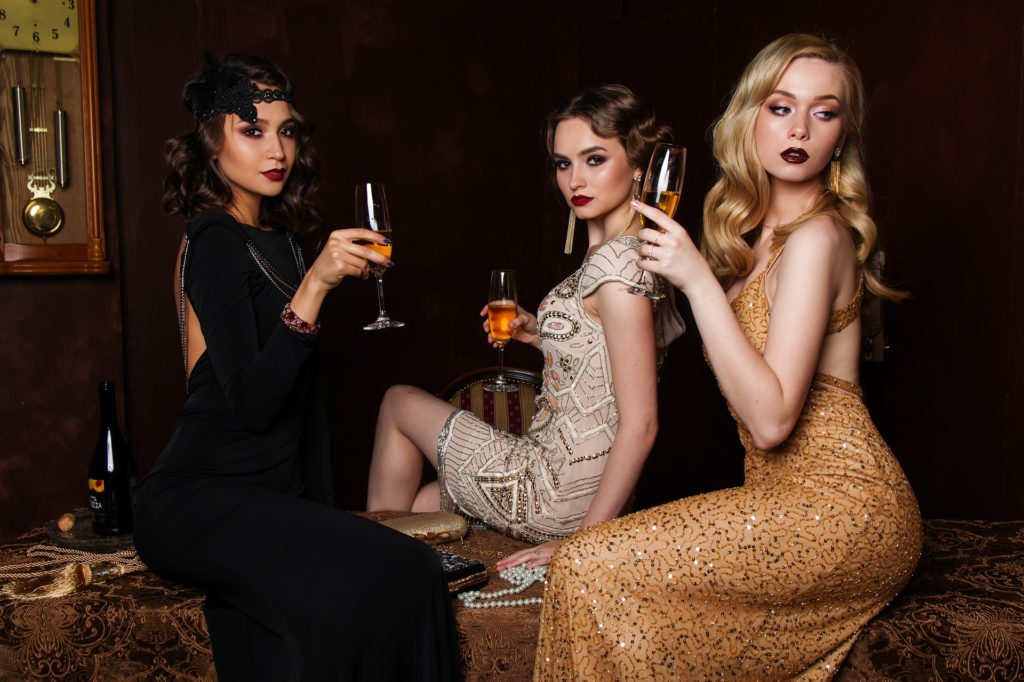 Ce sont trois femmes très apprêtées en robe de soirée qui boivent une coupe de champagne. 